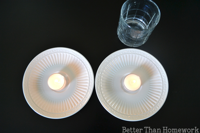 Two tealight candles that have been lit sitting up saucers with a clear drinking glass nearby